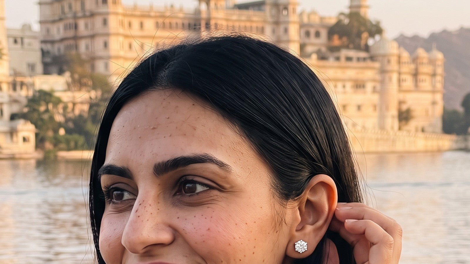 A woman tucking her hair behind her ear to show a diamond stud, a lakeside palace behind her