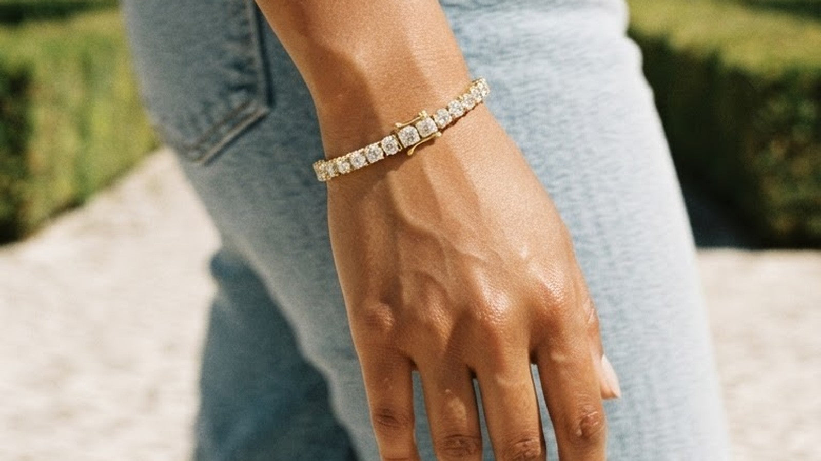 A diamond tennis bracelet on a woman's wrist as she walks, denim and garden path behind