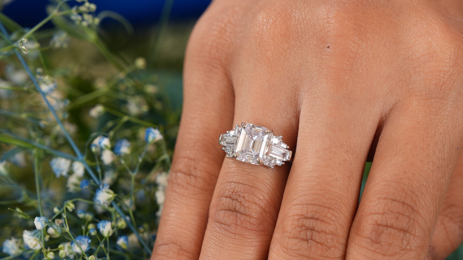 Emerald cut lab grown diamond ring with tapered side stones, worn on the hand among white flowers