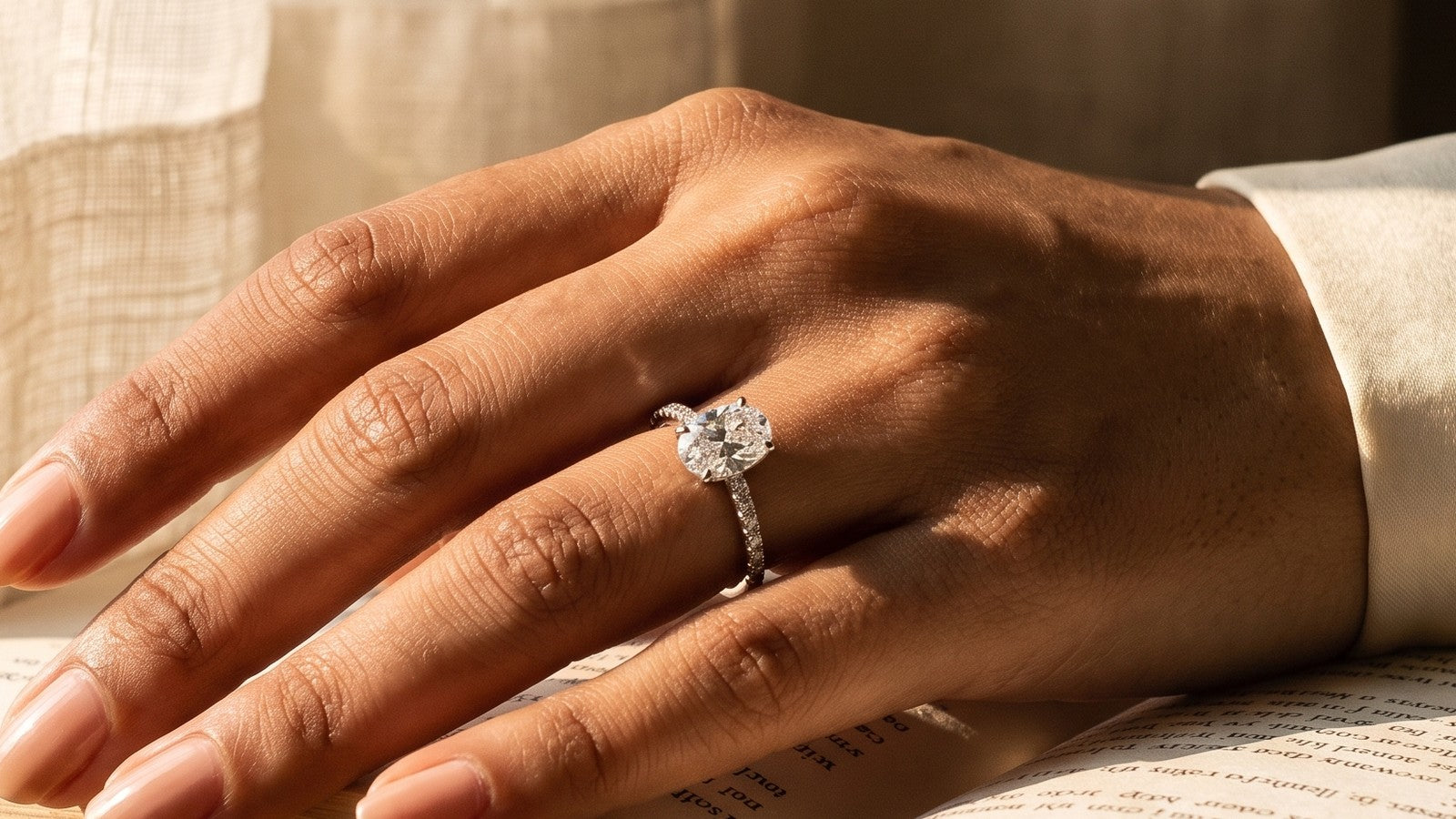 Oval lab grown diamond solitaire on a pave band, worn on a hand resting on an open book by the window