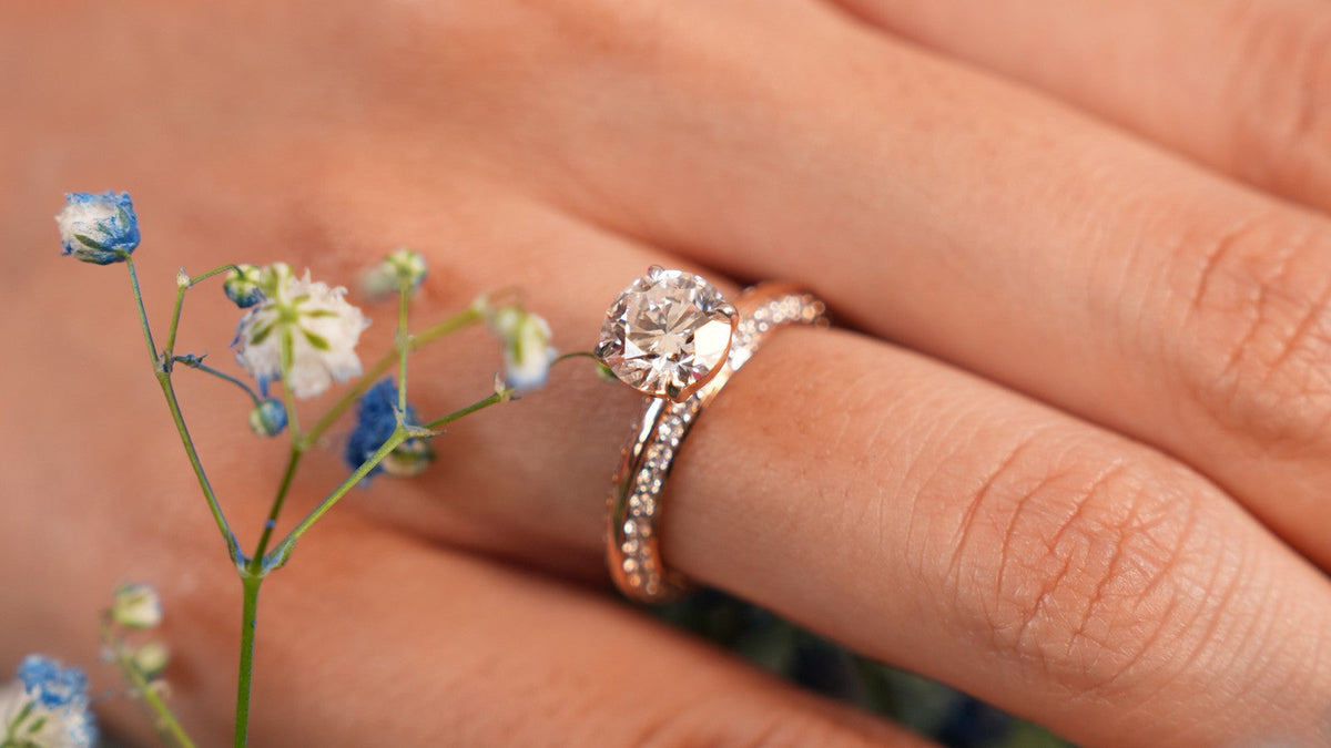 Round lab grown diamond solitaire on a rose gold pave band, worn on the hand beside small blue flowers